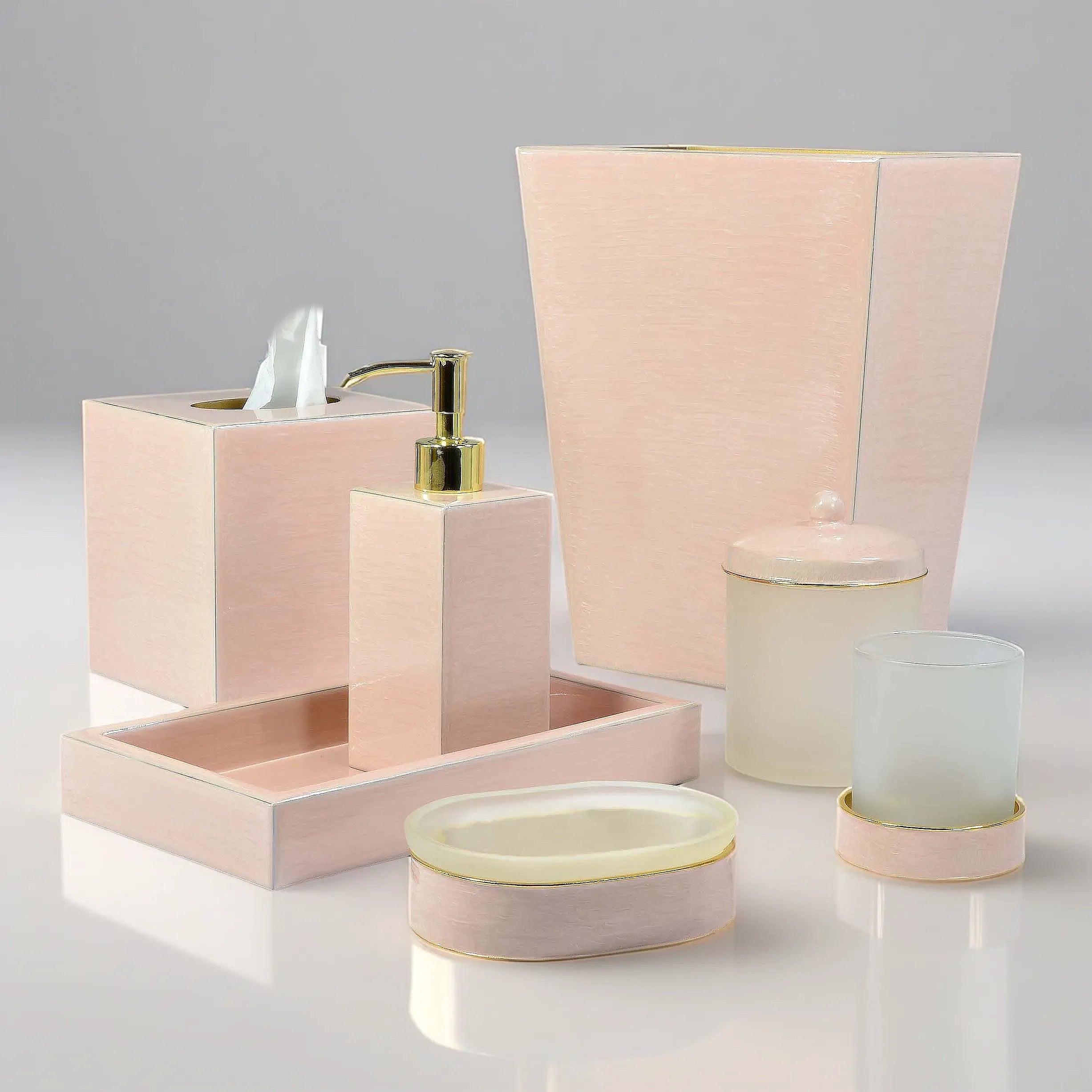 A coordinated bathroom or vanity accessory set in pale blush pink with small gold accents, including a tissue box holder, soap dispenser, tray, lidded container, round soap dish, and a frosted cup on a light gray studio background.